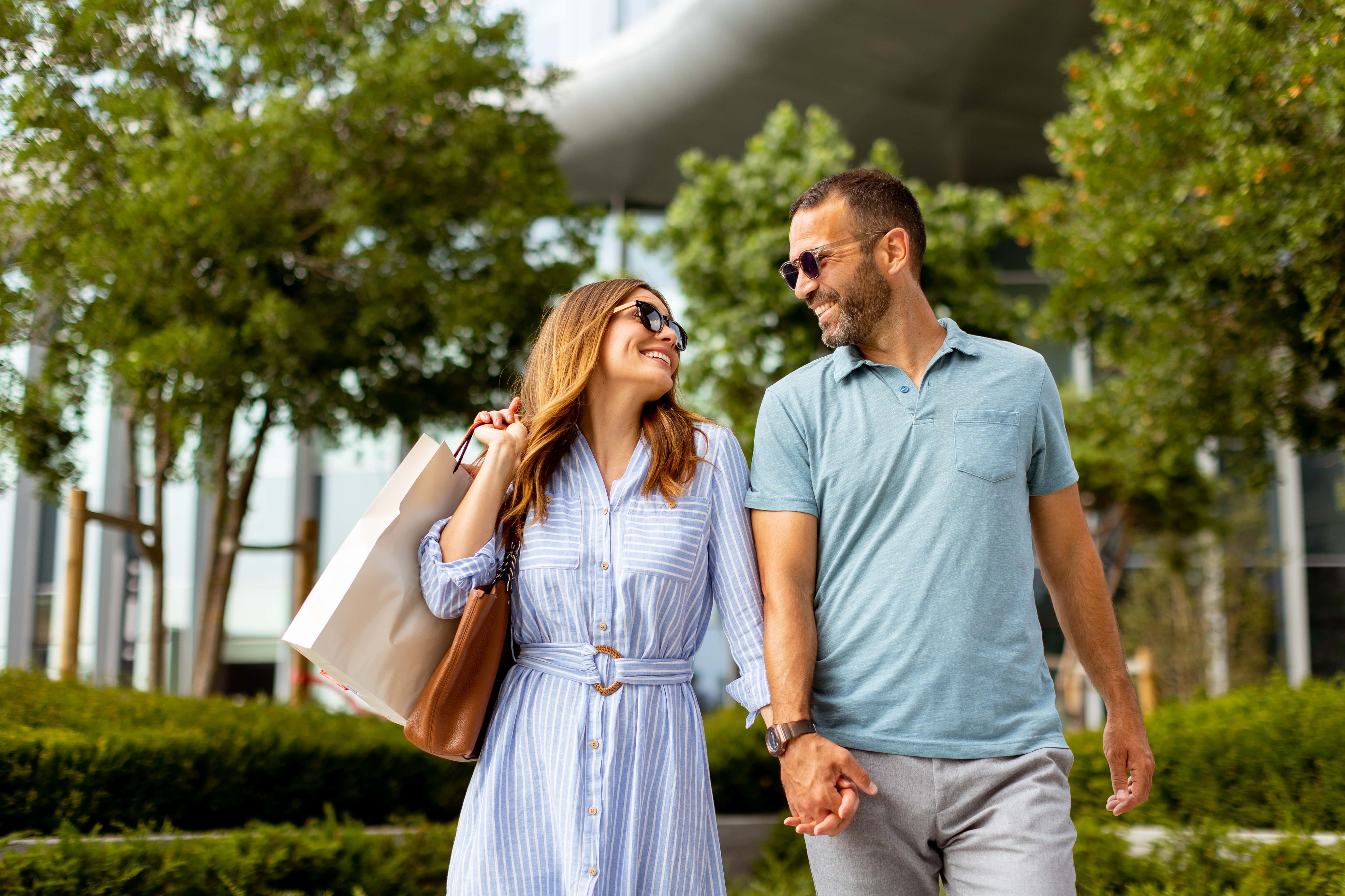 A couple walking hand in hand outdoors, smiling at each other while carrying shopping bags. The scene is surrounded by greenery and modern architecture in the background.