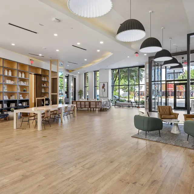 A spacious and modern lobby featuring wooden floors, a large bookshelf, seating areas with contemporary furniture, and large windows letting in natural light.