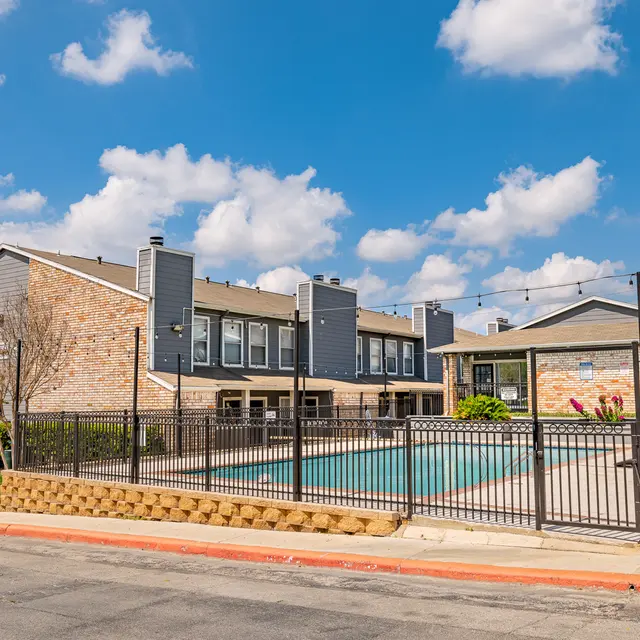Residential complex with a swimming pool surrounded by a fence under a blue sky with clouds.