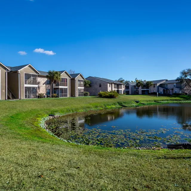 A scenic view of an apartment complex beside a pond, with green grass and clear blue skies.