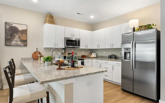 A modern kitchen with white cabinets, stainless steel appliances, and a granite countertop. The kitchen includes a breakfast bar with a few stools and decorative elements such as bottles and plants.