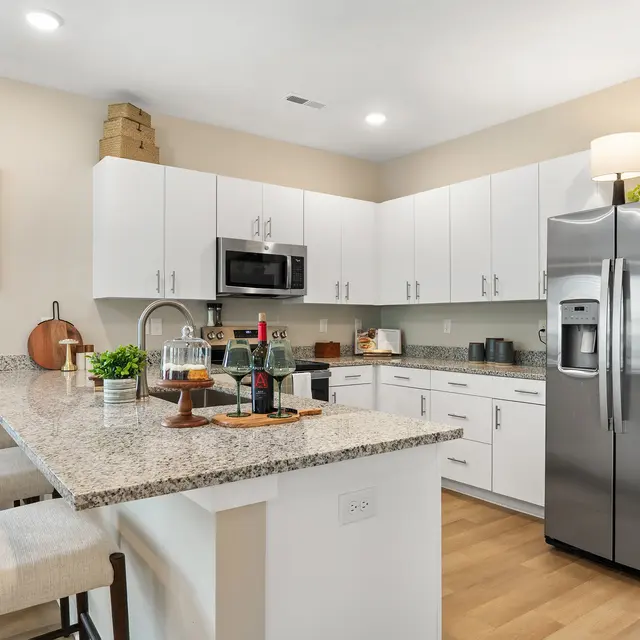 A modern kitchen with white cabinets, stainless steel appliances, and a granite countertop. The kitchen includes a breakfast bar with a few stools and decorative elements such as bottles and plants.