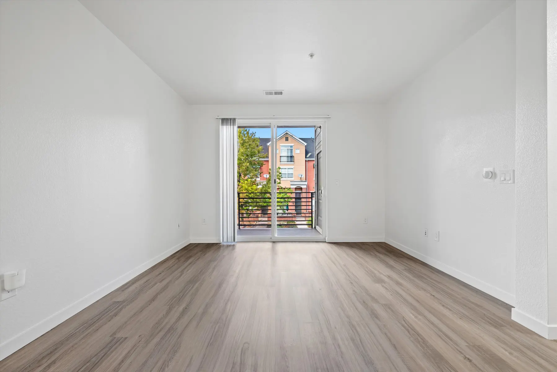 A spacious, empty living room with light-colored walls and a wooden floor, featuring a large sliding glass door that opens to a balcony with a view of greenery and nearby buildings.