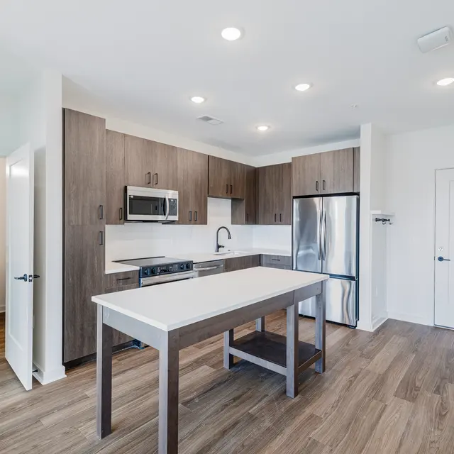 A modern kitchen with wooden cabinets and a central island. Stainless steel appliances are visible, along with a double sink. The kitchen features an open layout with good natural lighting.