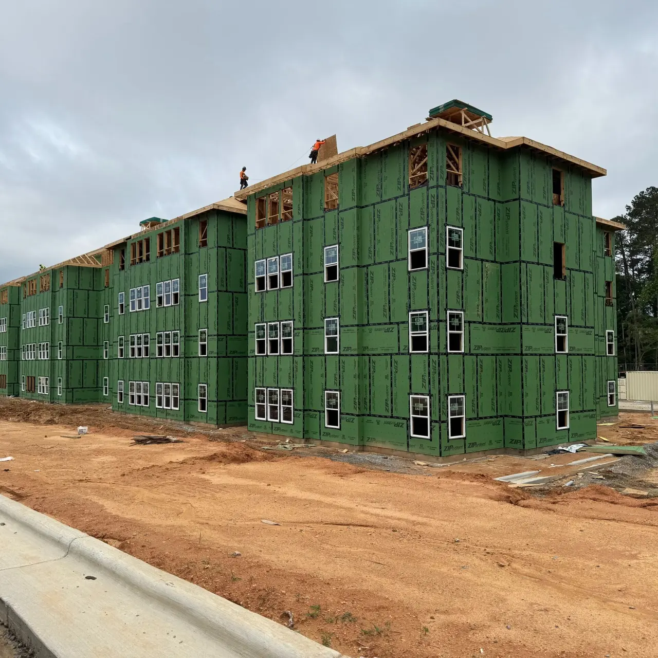 A partially constructed multi-story building with green sheathing and multiple windows on the facade, situated on a dirt lot with construction debris. Workers are visible on the roof in the background.