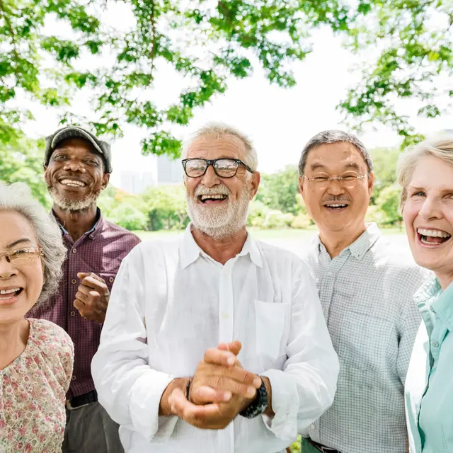 A joyful group of five seniors smiling and laughing together outdoors under a lush green tree canopy.