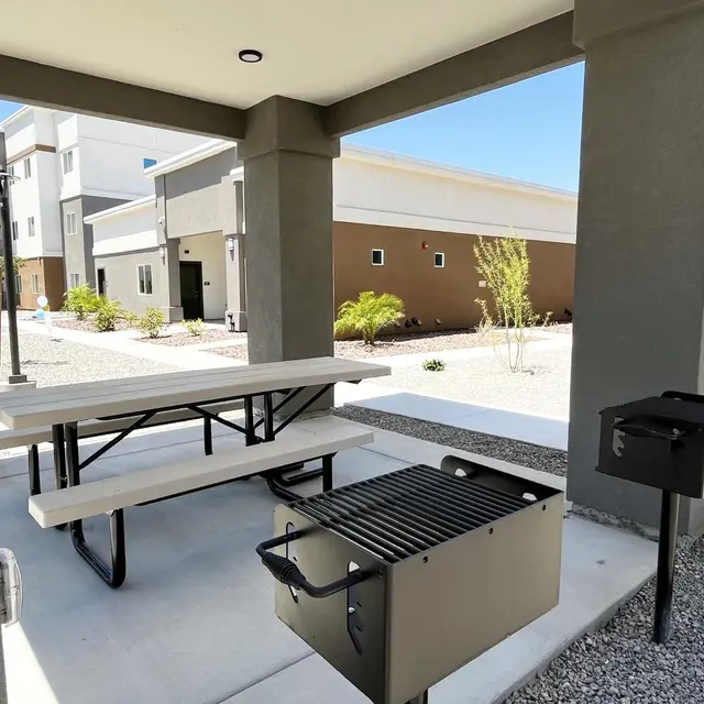 Outdoor Grill Area A shaded outdoor area featuring a picnic table, a grill, and a trash bin, surrounded by a gravel landscape and modern buildings in the background.