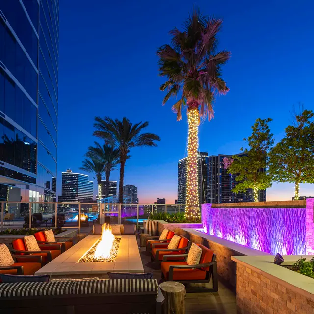 Modern Rooftop Lounge at Twilight A rooftop lounge area featuring a modern fire pit surrounded by comfortable seating, palm trees, and a water feature with illuminated purple lights, set against a city skyline at twilight.