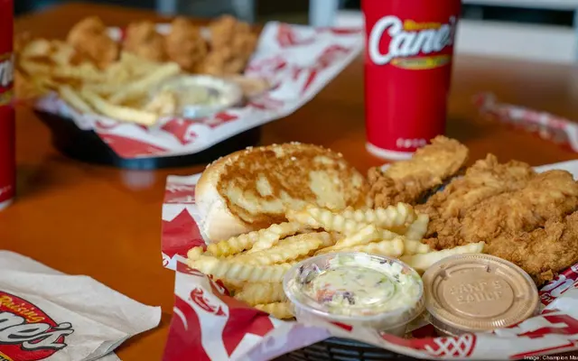 A delicious meal featuring fried chicken, crinkle-cut fries, coleslaw, and dipping sauces, set on a tray at a restaurant table with drinks in the background.