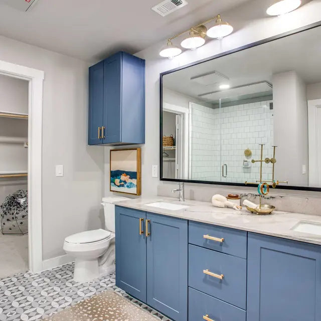 A modern bathroom featuring a double vanity with blue cabinetry, a large mirror, and a stylish light fixture. A toilet is positioned nearby, with a walk-in shower visible in the background. There is a walk-in closet adjacent to the bathroom, with a wooden shelf and some bags on it.