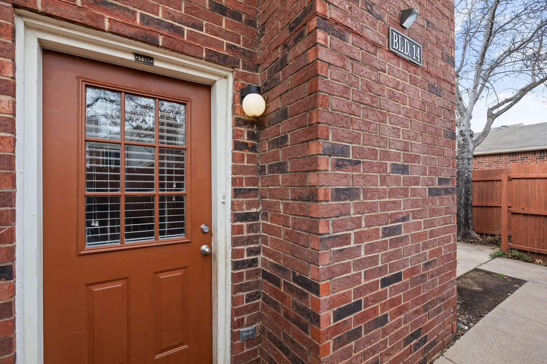 Close-up view of a brown door with grid window, leading into a brick building. A light fixture is mounted next to the door, and a sign reads 'BUILDING 11'.