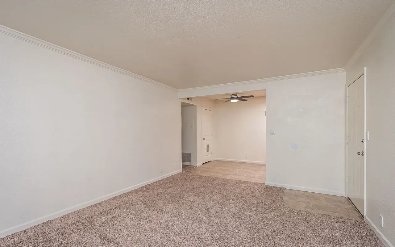 An empty living room with beige carpet and white walls, featuring a doorway leading to another room and a ceiling fan visible in the background.