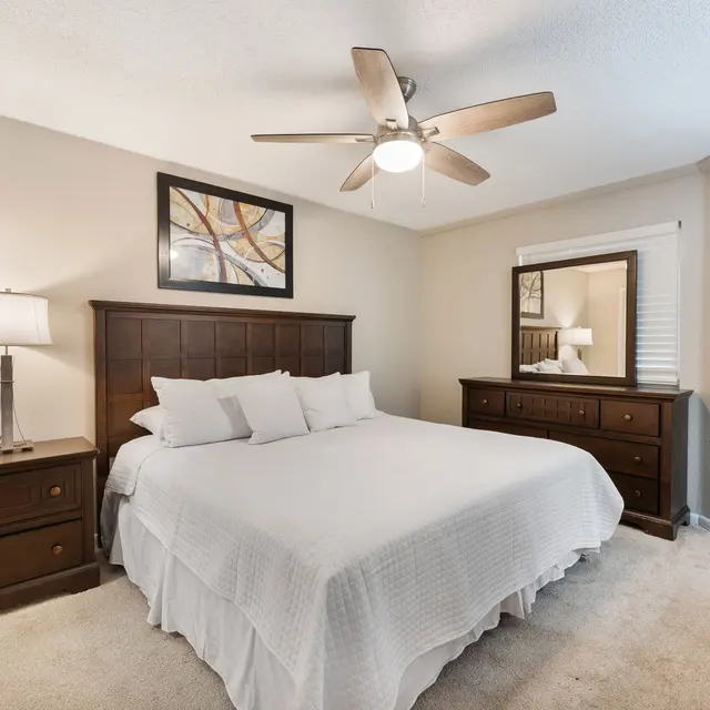 A well-furnished bedroom featuring a large bed with white linens, a bedside table with a lamp, a wooden dresser, and a mirror. The decor includes a framed artwork on the wall, with warm lighting and a ceiling fan.