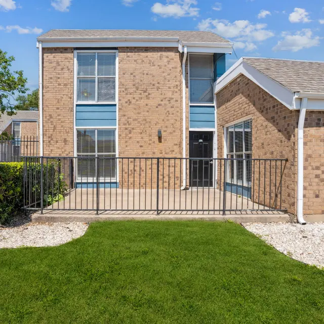 Exterior view of a brick apartment building with a fenced patio area and green lawn