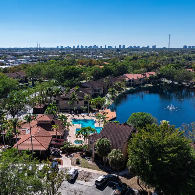 Aerial view of a residential area with a pool, surrounded by lush greenery and a lake in the foreground, with a city skyline in the distance under a clear blue sky.