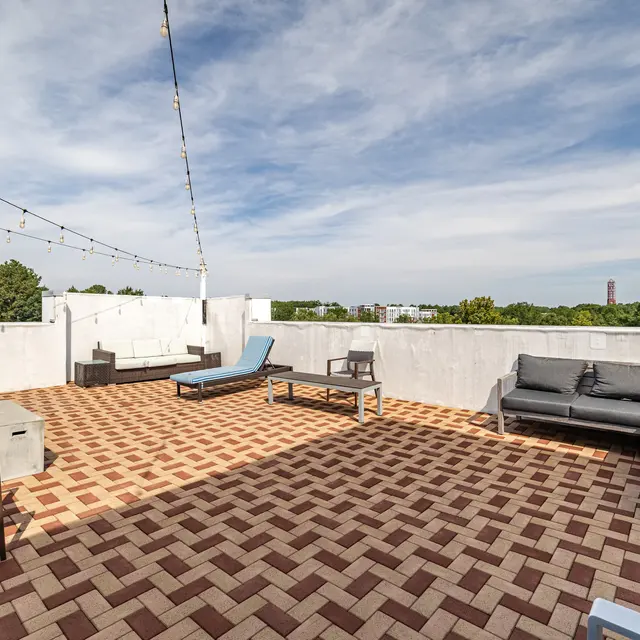 A spacious rooftop terrace featuring patterned tiled flooring, lounge chairs, and a sectional sofa. There's a clear sky in the background with some greenery visible.