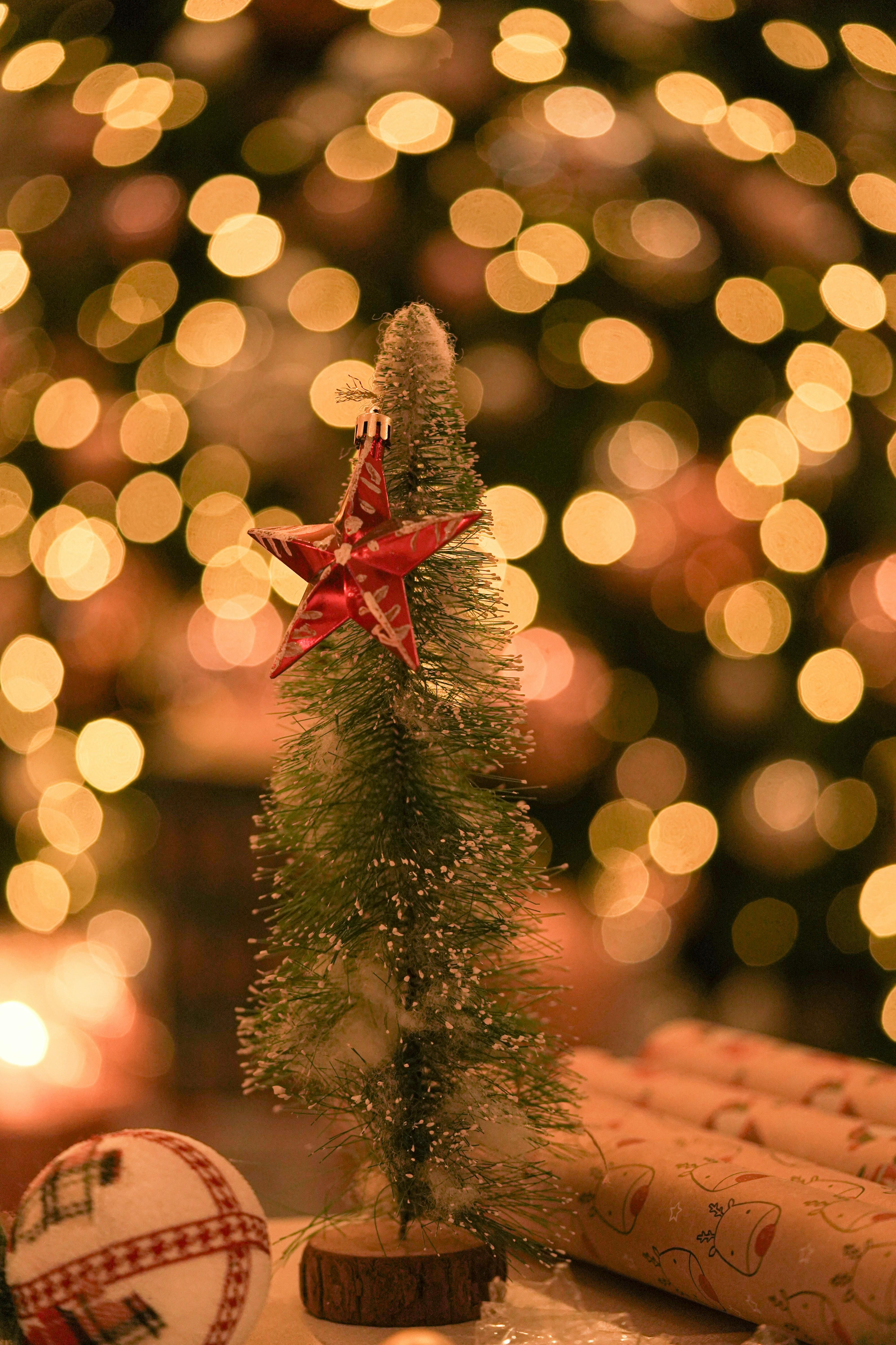 A small decorative Christmas tree with a red star on top, set against a backdrop of blurred golden lights. A round ornament and wrapped gifts are also visible in the foreground.