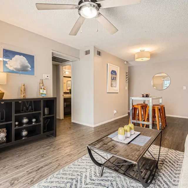 A cozy living room with a light-colored couch, a coffee table, and a small dining table in the background. There is a ceiling fan, a bookshelf with decorative items, and wooden flooring. A doorway leads to another room, and a round mirror is visible on the wall.
