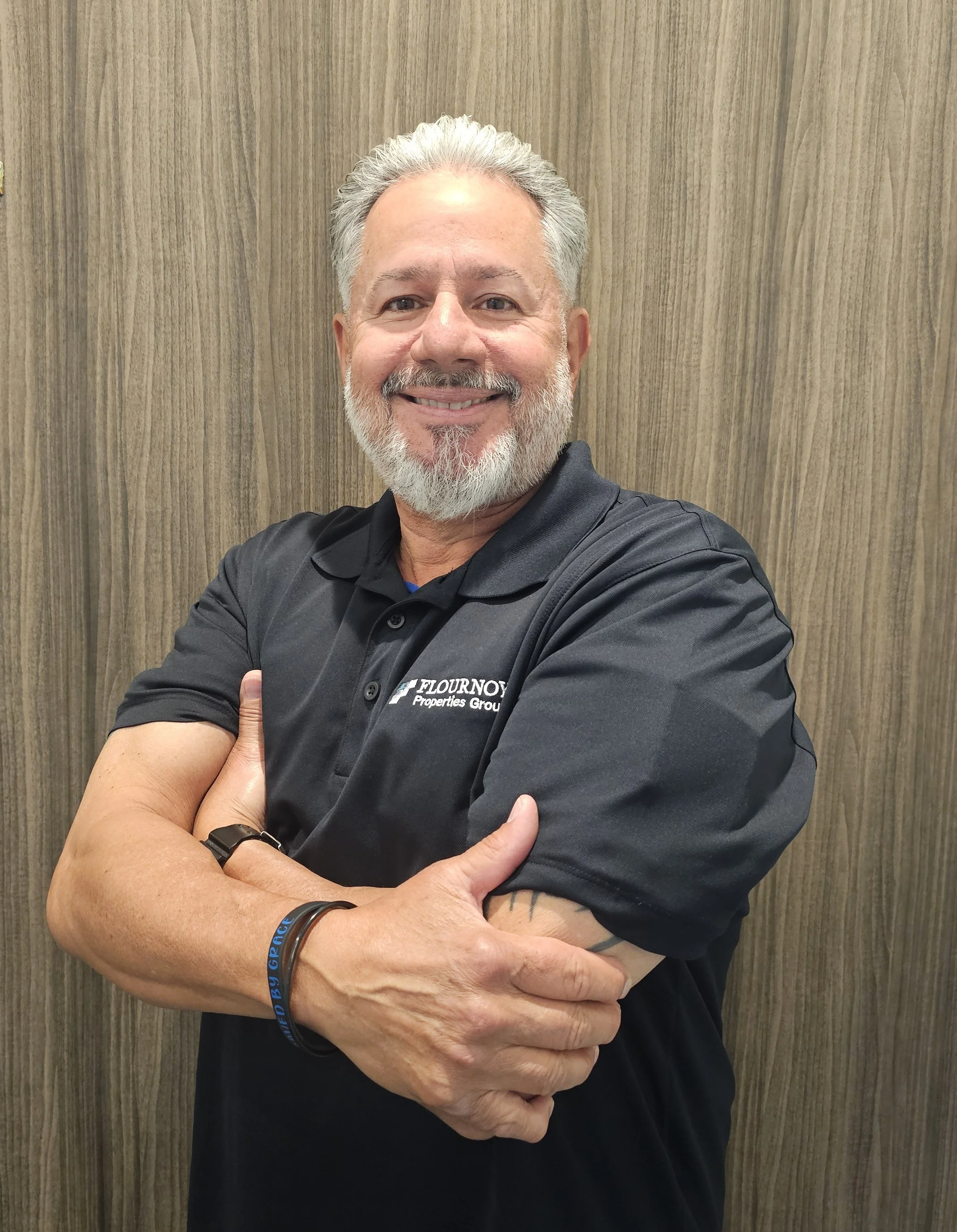 A smiling man with gray hair and a beard, wearing a black polo shirt with a logo. He is standing with his arms crossed in front of a wooden background.