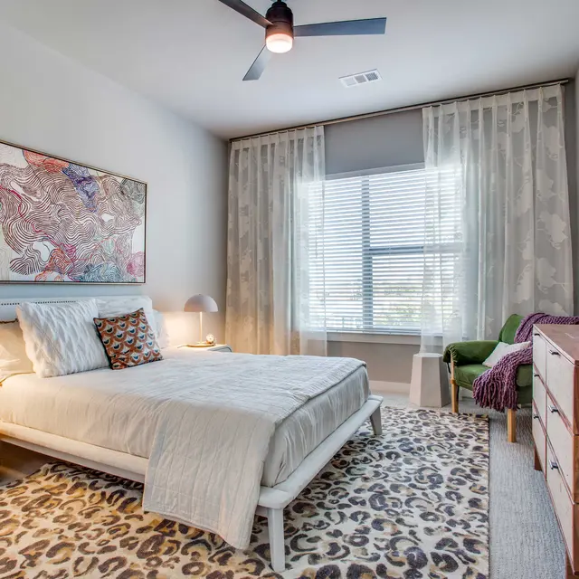 A modern bedroom featuring a white bed with decorative pillows, a wooden nightstand with a bedside lamp, a large window with sheer curtains, a green accent chair, and a wooden dresser. The floor is covered with a patterned rug and there is a colorful wall art piece above the bed.