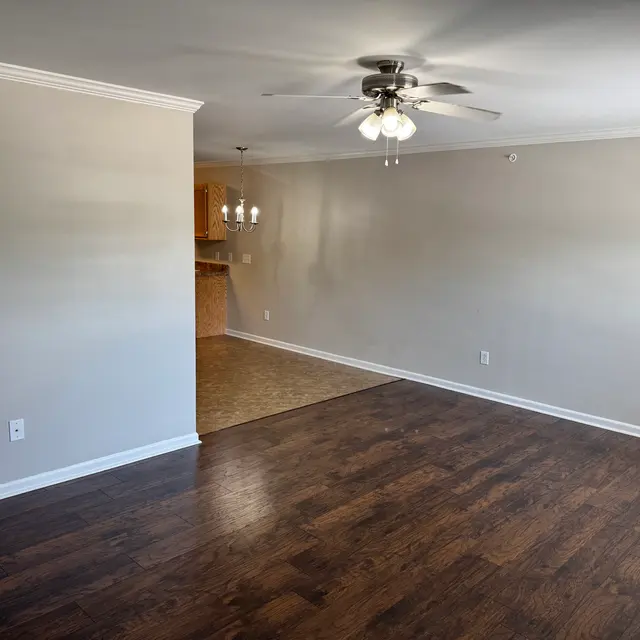 Spacious Living Area with Modern Design A spacious living area featuring brown wooden flooring, a ceiling fan, and light beige walls. There is an open space leading to a kitchen area with light wood cabinets, and a small dining area with a chandelier.