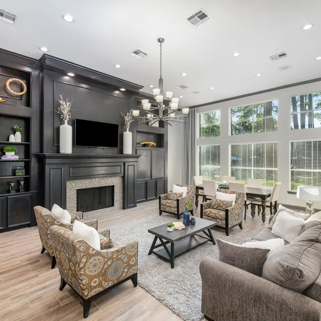 A modern living room with black cabinetry, a chandelier, and large windows overlooking a wooded area. The room features comfortable seating arrangements, including stylish armchairs and a plush sofa, as well as decorative elements on shelves.