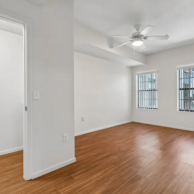 Empty Apartment Room An empty room with wooden flooring, a ceiling fan, and three windows with metal bars. A doorway leads to another room on the left.