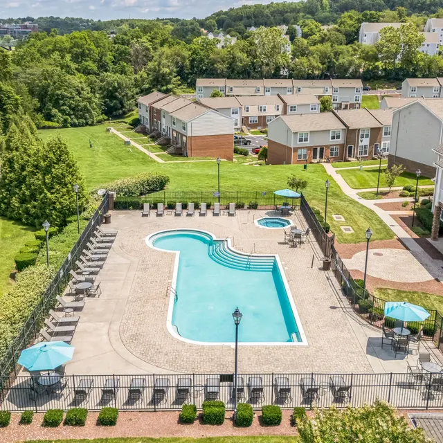 Aerial View of Apartment Complex Pool Area Aerial view of a residential complex featuring a swimming pool with lounge chairs and umbrellas, surrounded by greenery and townhouses in the background.