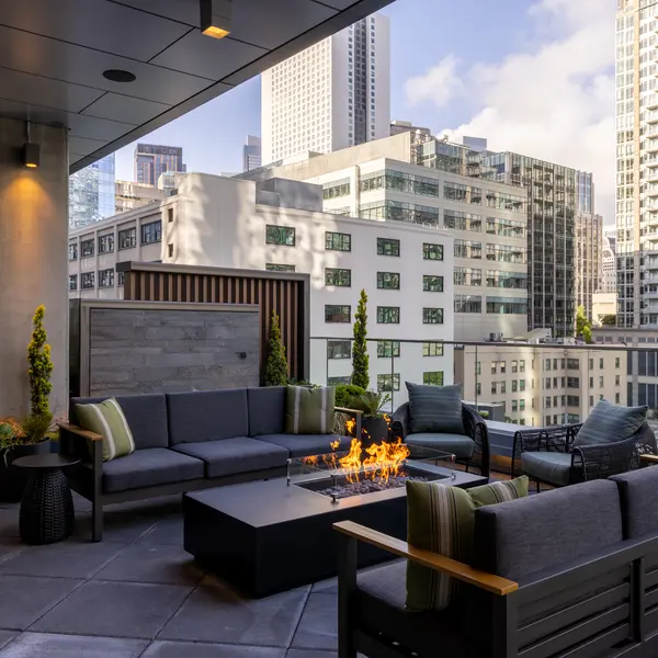 An outdoor lounge area featuring modern seating arrangements and a fire pit, with tall city buildings in the background.
