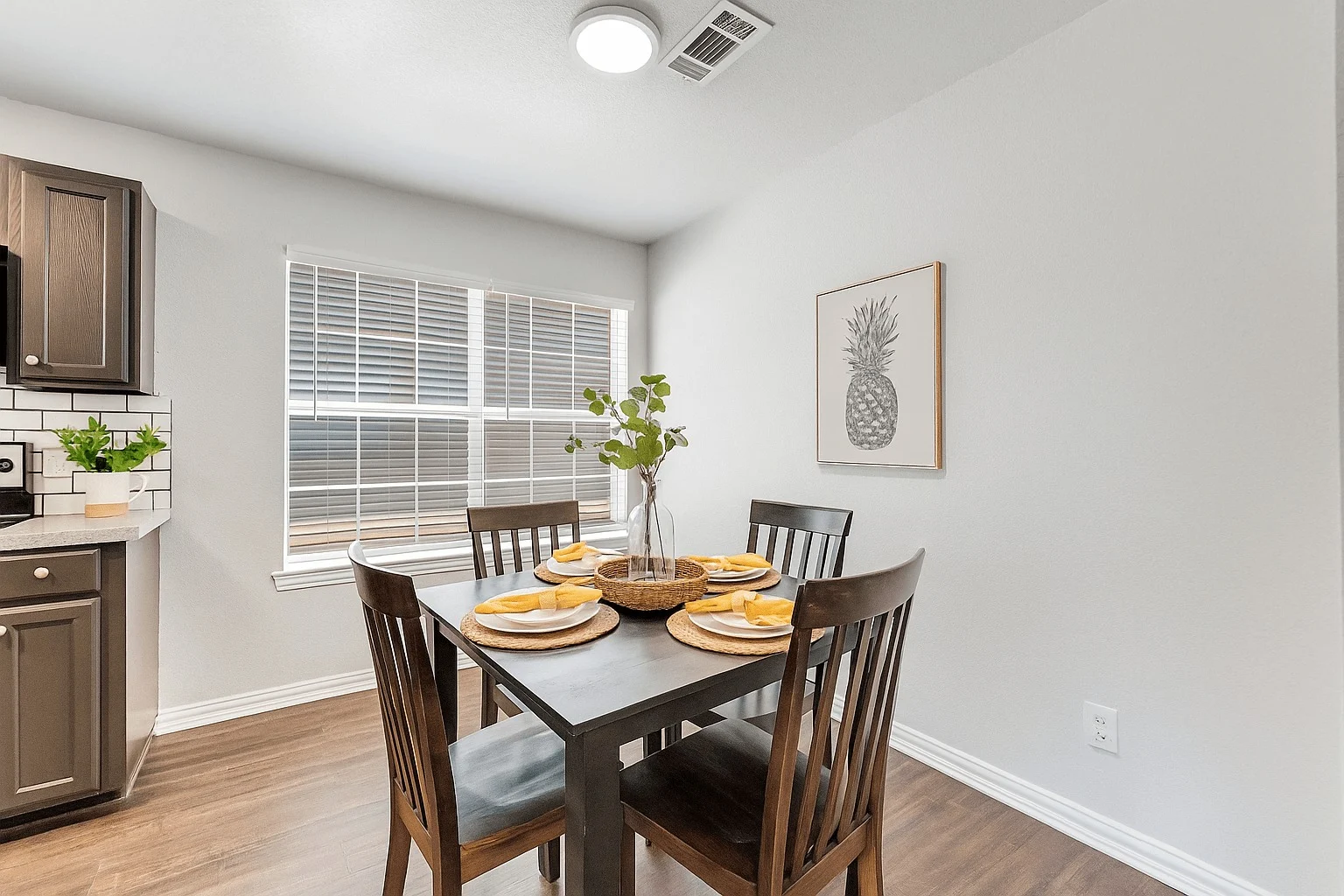 A modern dining area featuring a square table with four wooden chairs, set with yellow plates. A vase of greenery sits in the center of the table. In the background, there is a kitchen area with dark cabinets and a window with blinds. A pineapple artwork is on the wall.