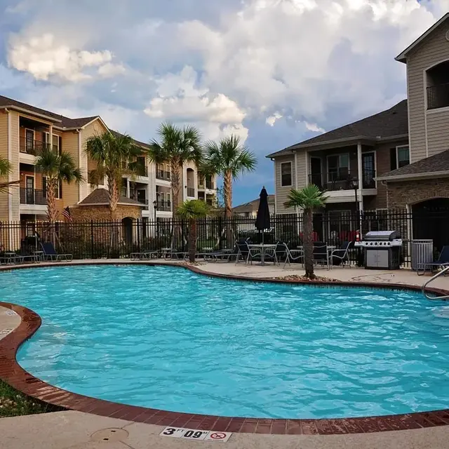 A swimming pool surrounded by palm trees and lounge chairs in an apartment complex. The sky is partly cloudy, and there are multiple buildings in the background.