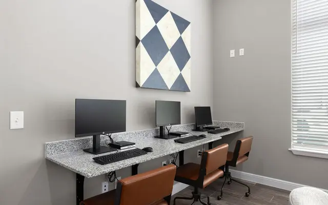 A modern shared computer area featuring four desktop computers on a long countertop, with brown ergonomic chairs, and a geometric wall art piece above in blue and cream colors.