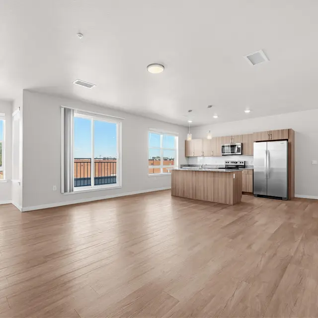 A spacious, modern apartment interior with a wooden floor, large windows, and a kitchen area featuring stainless steel appliances.