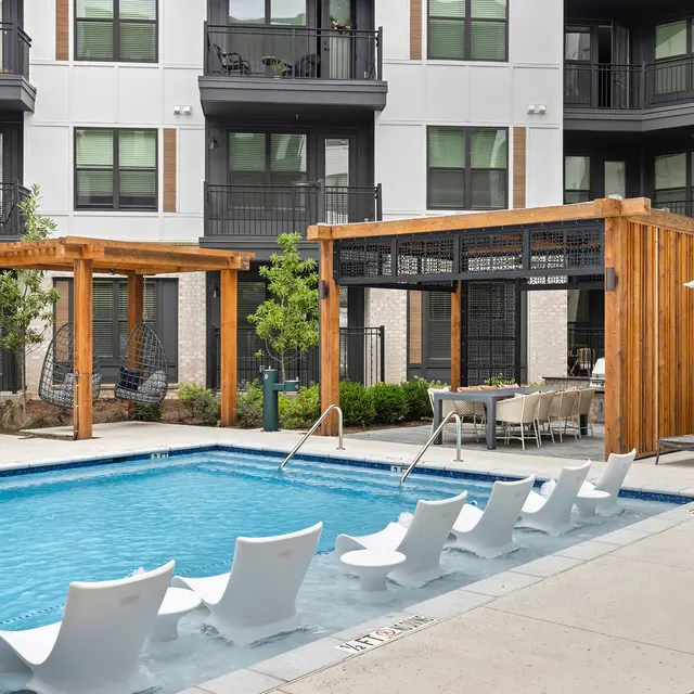 A modern apartment complex featuring a pool area with white lounge chairs and wooden pergolas, surrounded by greenery and contemporary buildings.