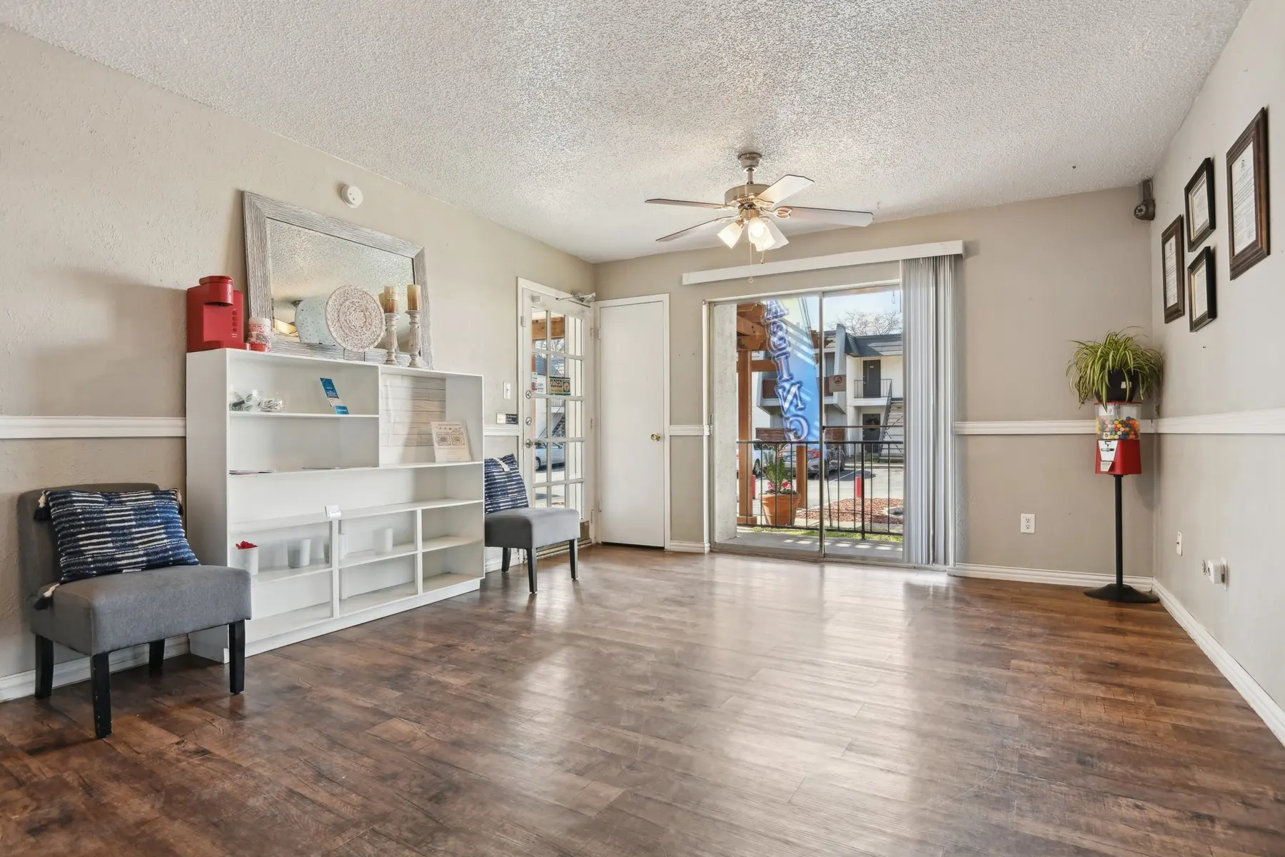 A spacious and bright living room featuring a small shelf, two chairs, a ceiling fan, and sliding glass doors leading to an outdoor area. The floor is wooden, and there are decorative pieces on the shelf and walls.