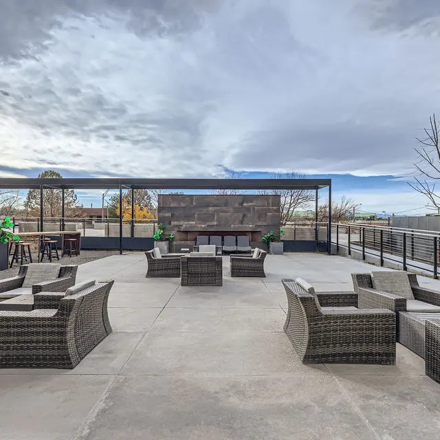 A spacious rooftop lounge area with comfortable wicker seating and a barbecue grill. The sky is partly cloudy and there are minimal trees in the background.