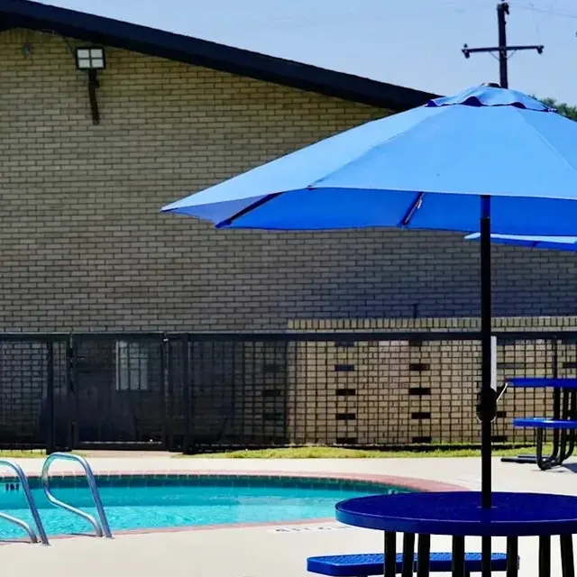 A swimming pool area with blue umbrellas and seating near a building. The pool is enclosed with a fence.