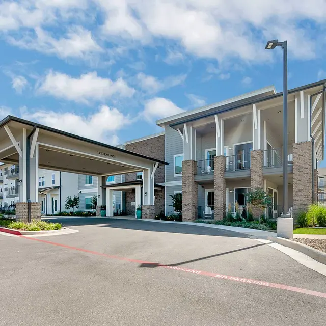 Modern apartment building with a covered entrance, landscaped garden, and blue sky in the background.