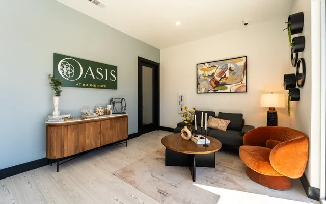 A modern lobby interior featuring a chic seating area with a gray sofa, orange accent chairs, and a coffee table. The decor includes a large wall sign that reads 'OASIS AT ROUND ROCK' and contemporary artwork on the wall. The space has light-colored flooring and natural lighting.
