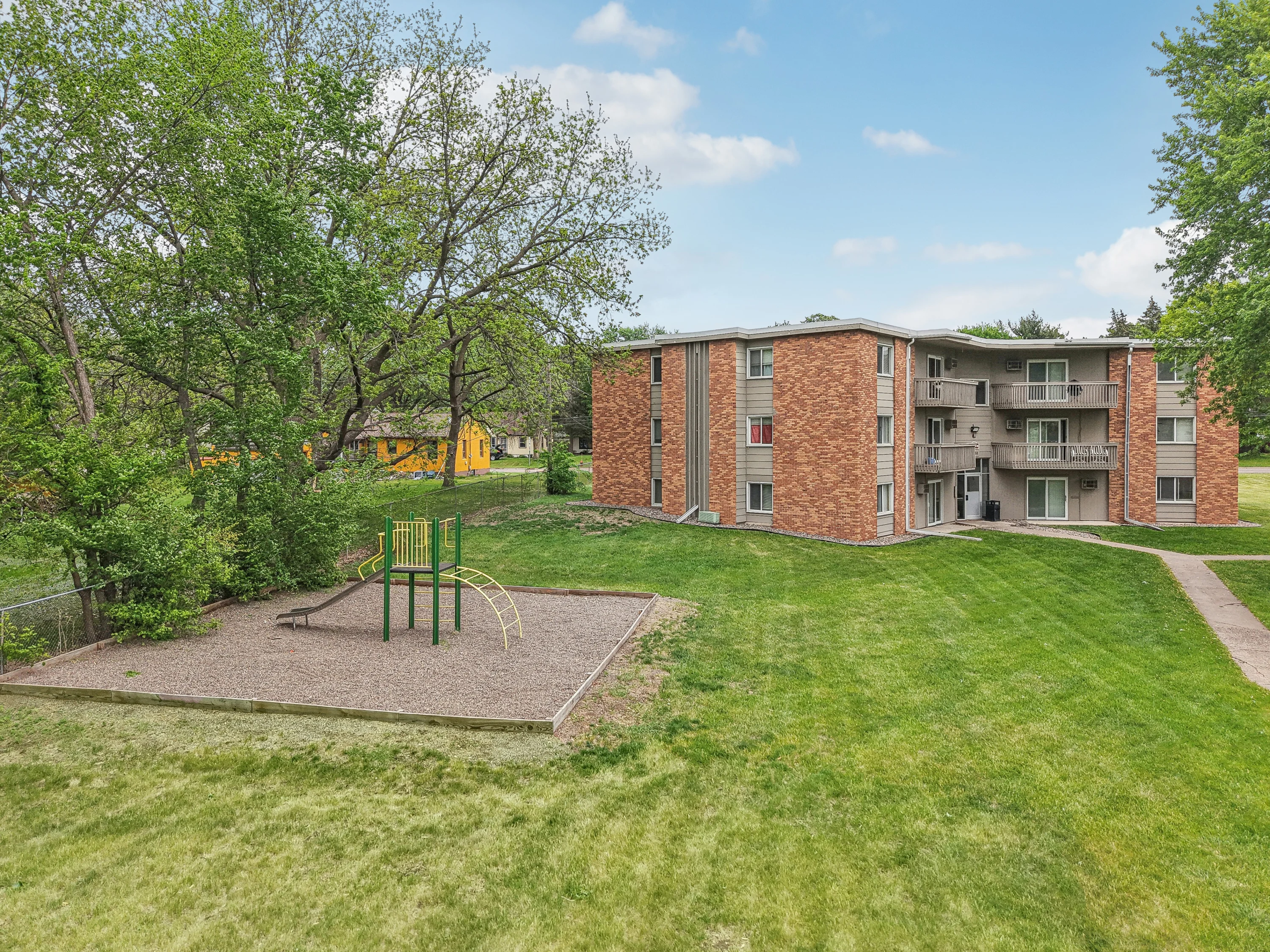 Apartment Community with Playground An outdoor view of a three-story brick apartment building with a green lawn and a children's playground featuring a slide and climbing structure.