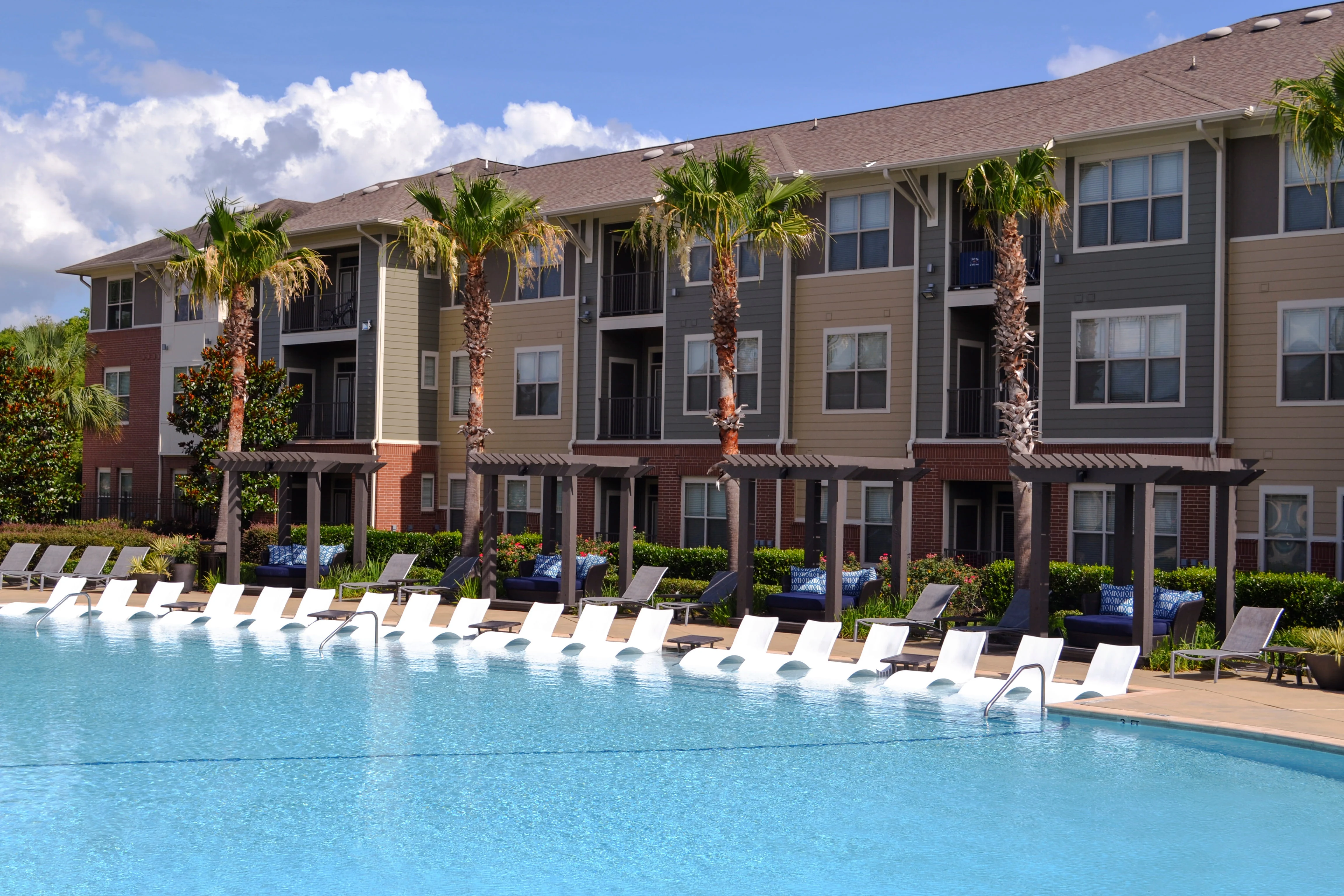 A bright pool area with sun loungers lined up beside a large swimming pool, surrounded by palm trees and an apartment building in the background.