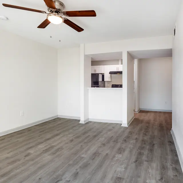 An empty living room featuring light-colored walls, a wooden floor, and a ceiling fan. The room opens to a kitchen area in the background with a view of some kitchen appliances.