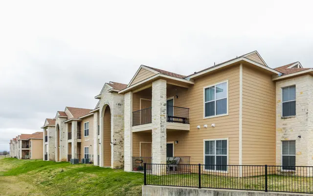 A row of modern apartment buildings featuring a mix of stone and siding exteriors, with balconies and large windows, set against a grassy area.
