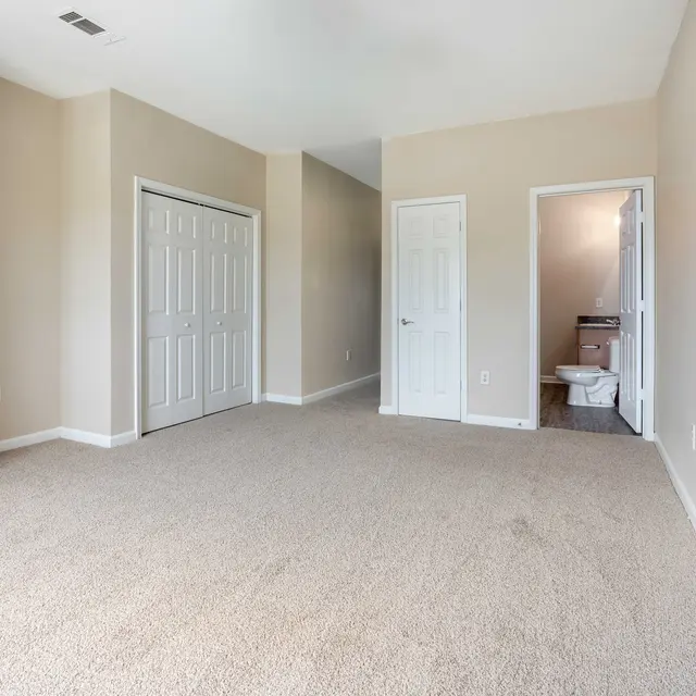A spacious empty room with beige carpet, featuring a closet with double doors and an entrance leading to a bathroom at the back.