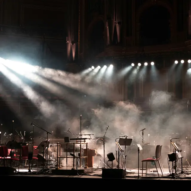 An empty concert stage with a variety of musical instruments and chairs, illuminated by beams of light. Fog fills the air, creating an atmospheric scene.