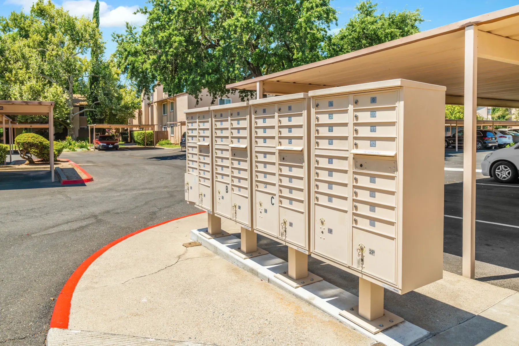 A row of community mailboxes situated outside an apartment complex, with trees and a parking area in the background.