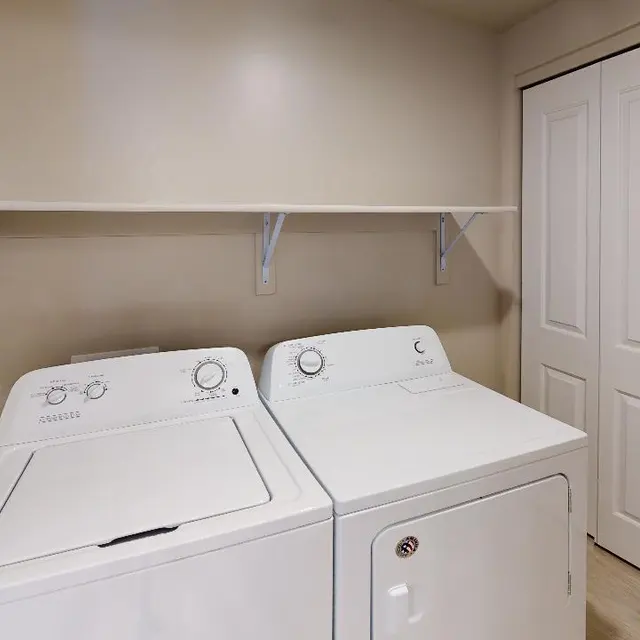 A laundry room featuring a washing machine and dryer side by side, with a shelf above them and closet doors in the background.