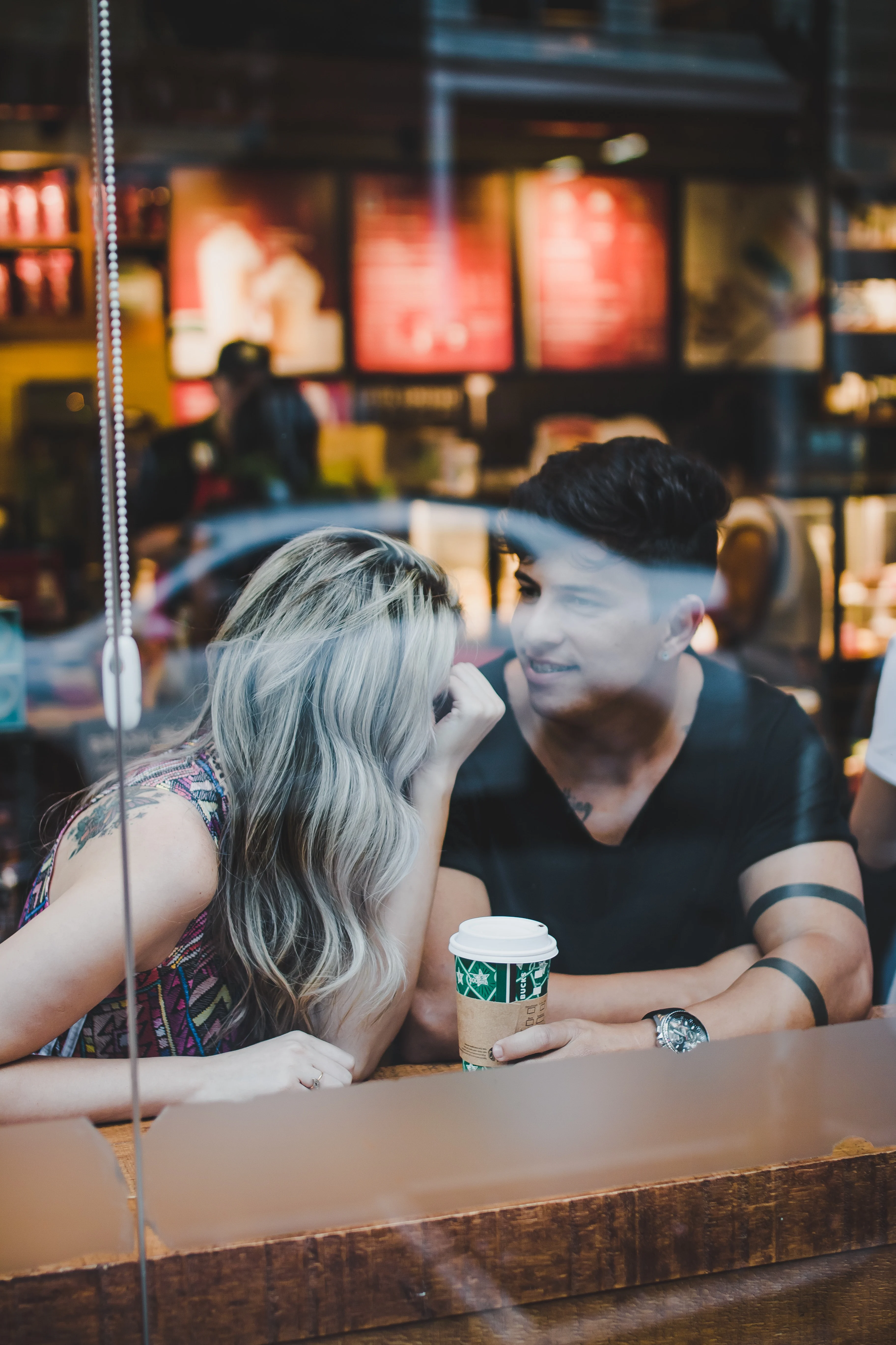 A couple sitting at a coffee shop, leaning close and sharing a smile, with a Starbucks cup on the table between them.