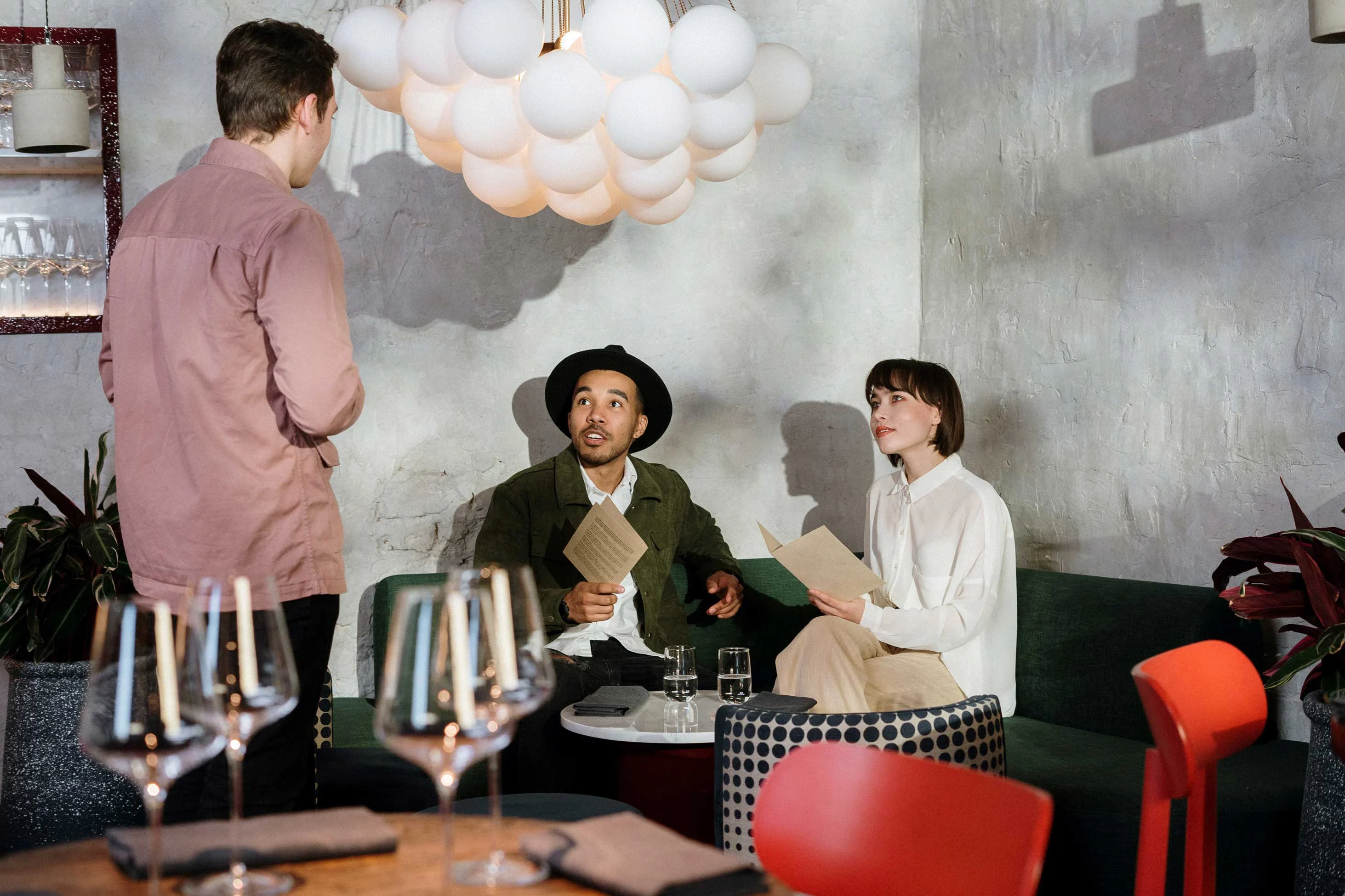 A waiter takes an order from a couple seated at a table in a stylish restaurant with unique lighting and decor.