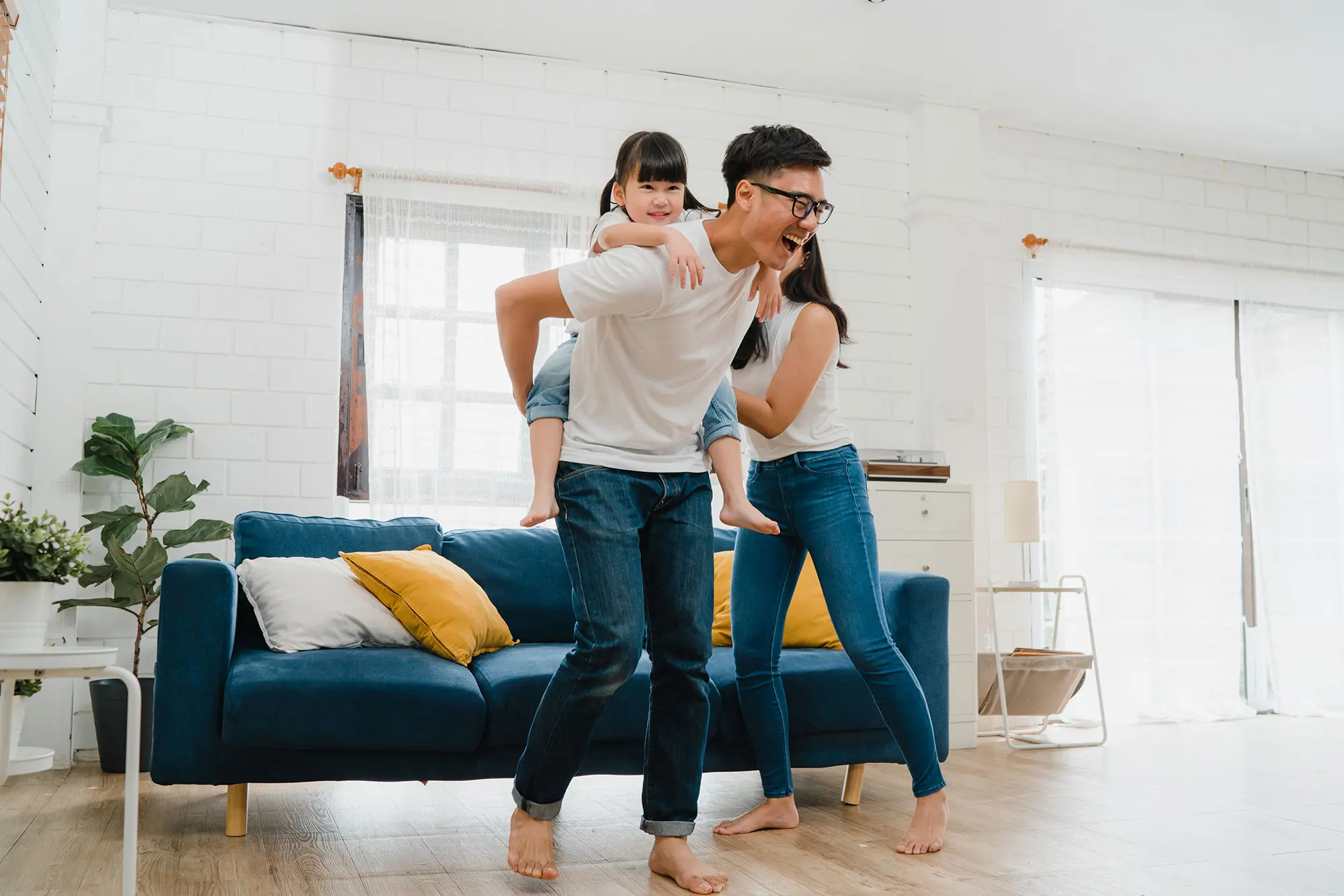 A family having fun in a living room; a man giving a piggyback ride to a child while a woman smiles next to a blue sofa.